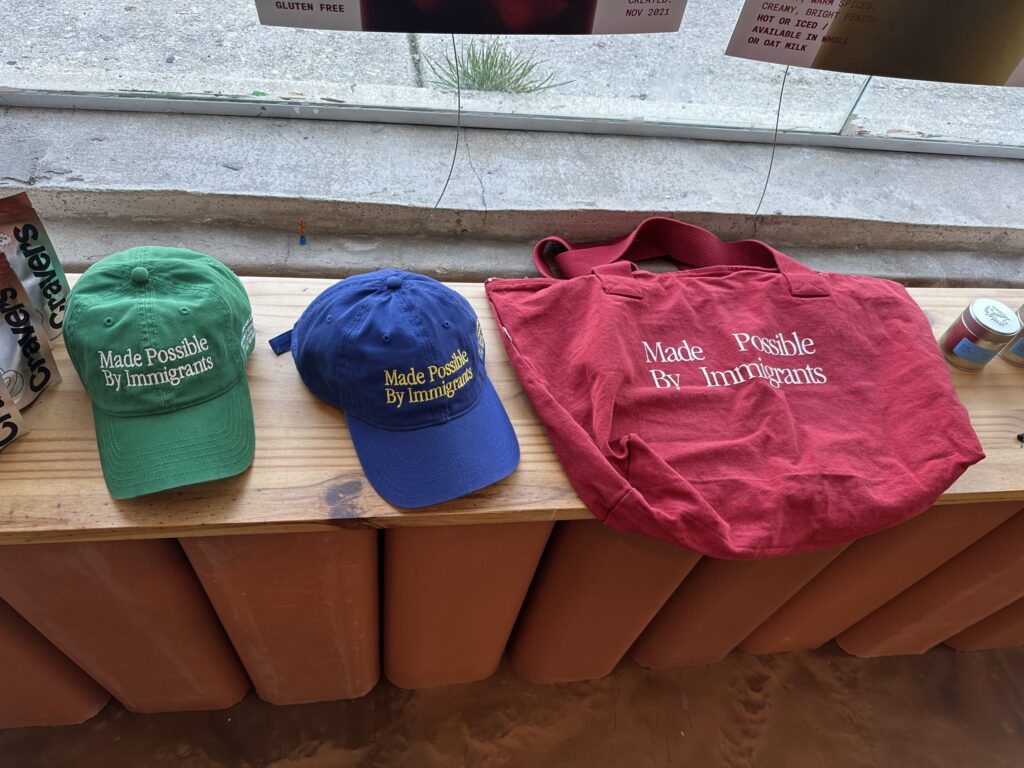 Caps and tote embroidered 'Made Possible By Immigrants' in the BuenoDays window