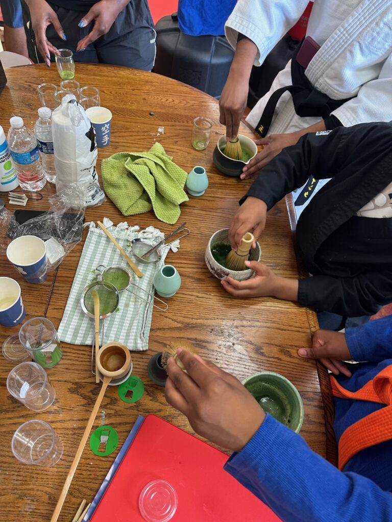 Hands whisking bowls of matcha around a shared table, martial arts uniforms visible
