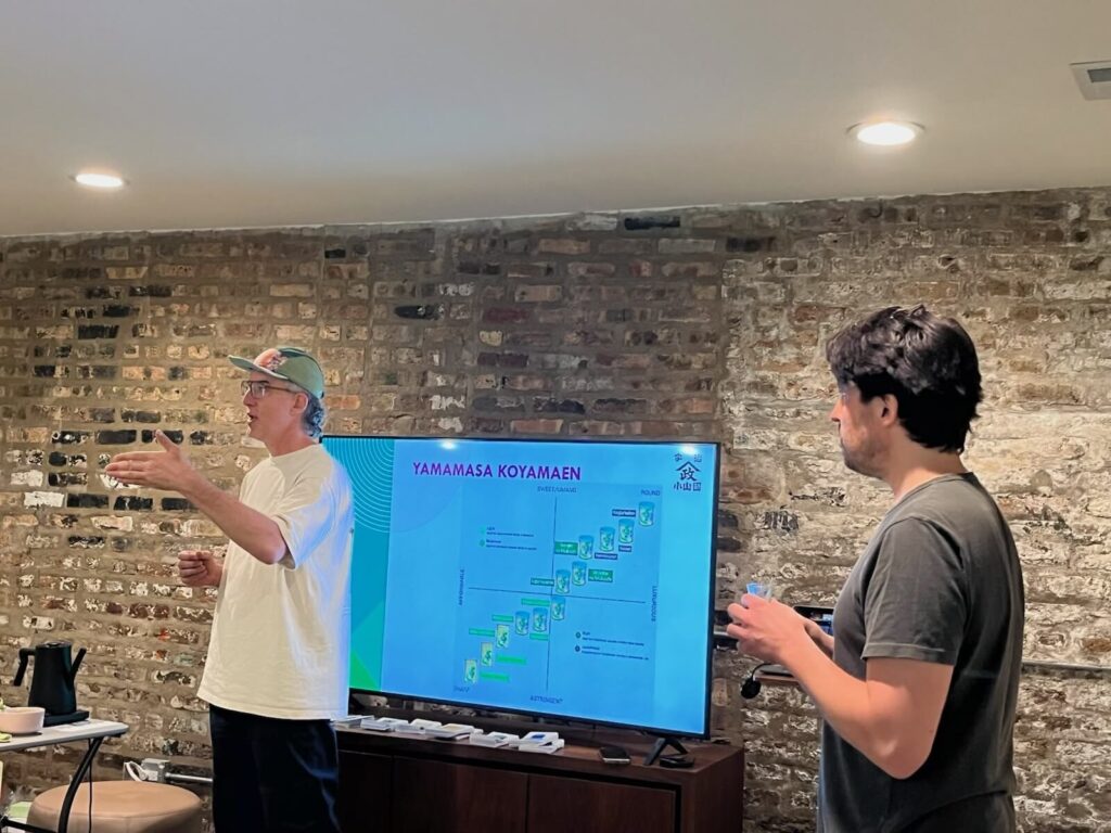 Two hosts beside a TV showing the Yamamasa Koyamaen slide, one of them talking to the room